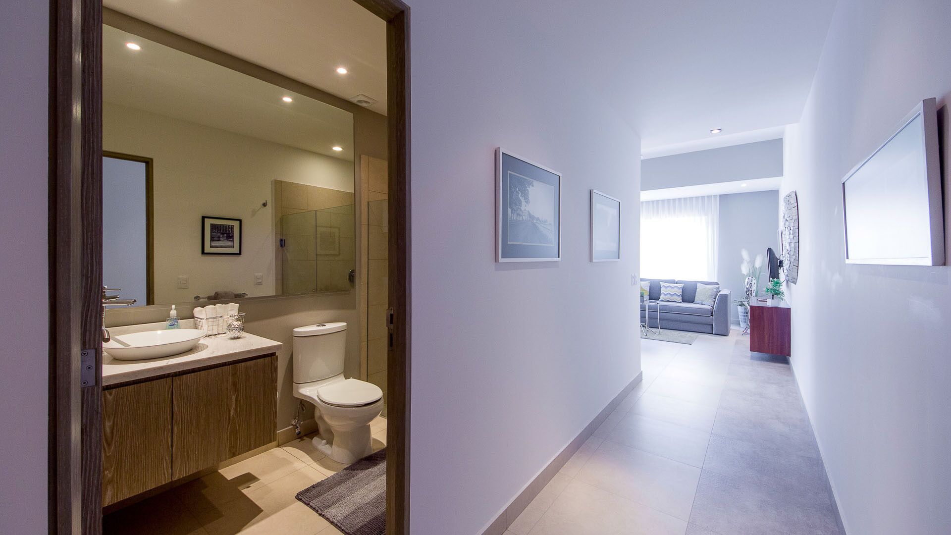 Une salle de bain moderne avec un grand miroir, un lavabo et des toilettes. Un couloir mène à un espace de vie comprenant un canapé et une table basse.
