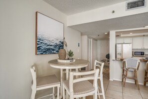 A dining area with a round table and four chairs, a kitchen with white cabinets and a microwave, and a large framed picture on the wall.