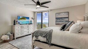 A bedroom with a bed, a ceiling fan, a dresser, a TV, and a large window with a view of the ocean. - Trillium 1B Completely remodeled, Beach Views from all rooms, top location! (Madeira Beach)