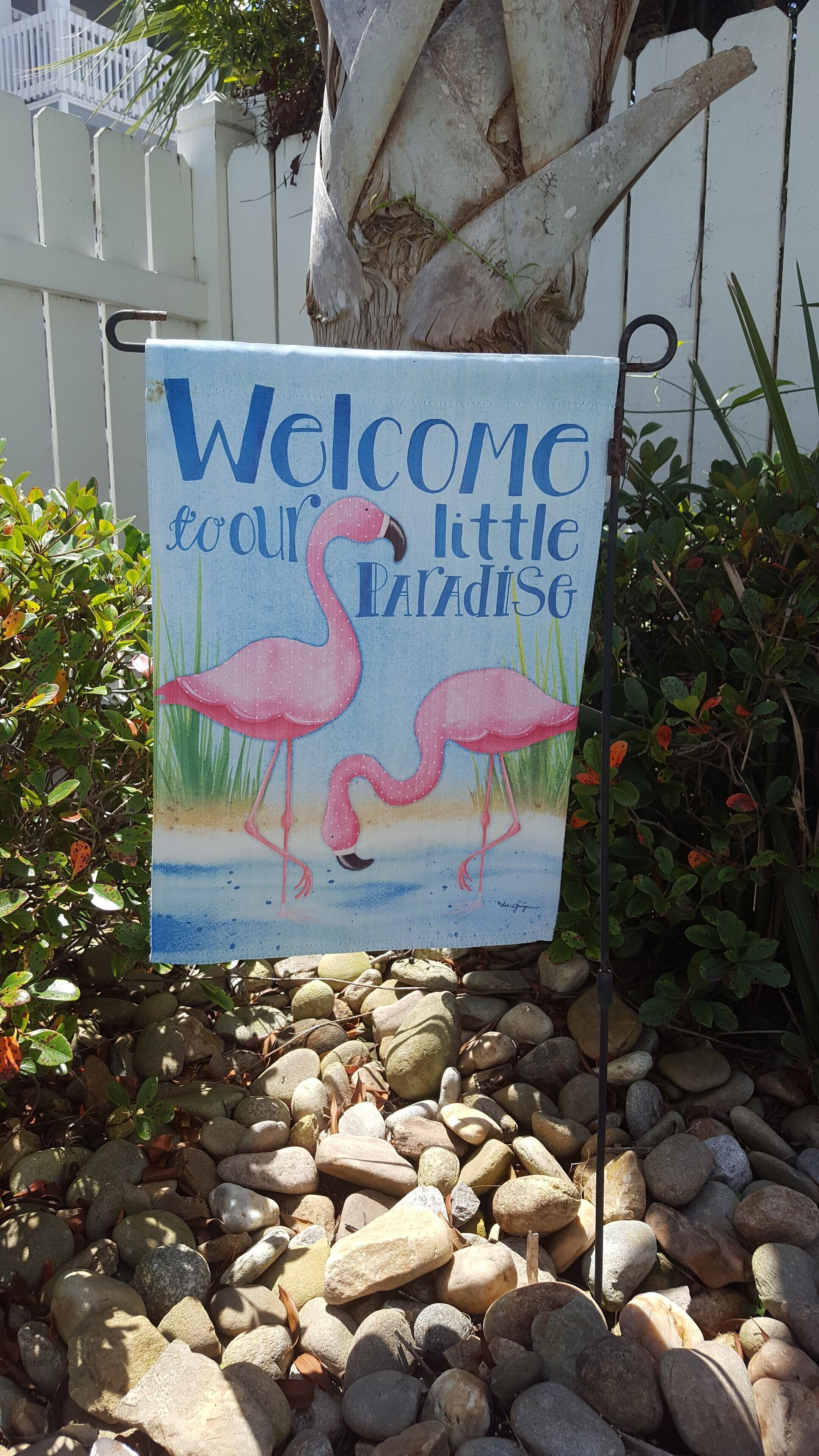 A welcome sign with flamingos and the text "Welcome to our little Paradise" in a garden setting with rocks and foliage.