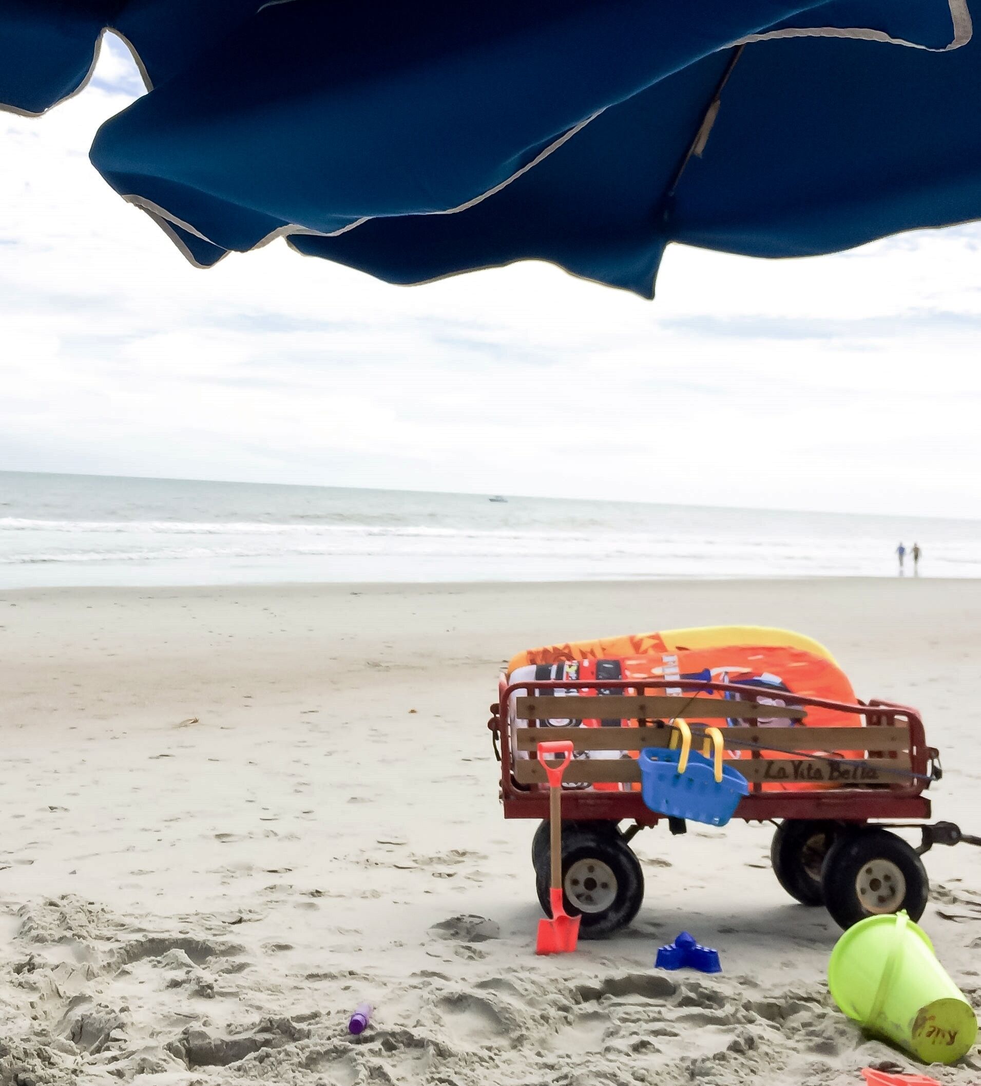 Escena de playa con un juguete a motor y juguetes para la arena bajo una sombrilla azul.