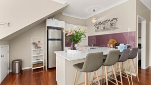 A modern kitchen with a central island, bar stools, and a decorative wall piece. - Husky Haven by Experience Jervis Bay (Huskisson)
