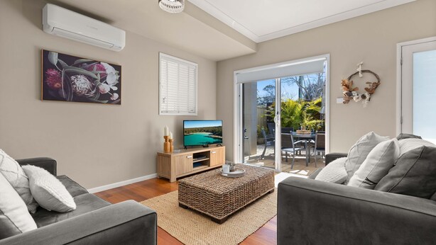 A modern living room with grey sofas, a TV on a wooden stand, a wicker ottoman, and a view of the outdoor patio. - Husky Haven by Experience Jervis Bay (Huskisson)