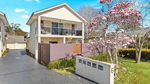 A modern two-story house with a balcony, a driveway, and a well-maintained garden with flowering trees. - Husky Haven by Experience Jervis Bay (Huskisson)