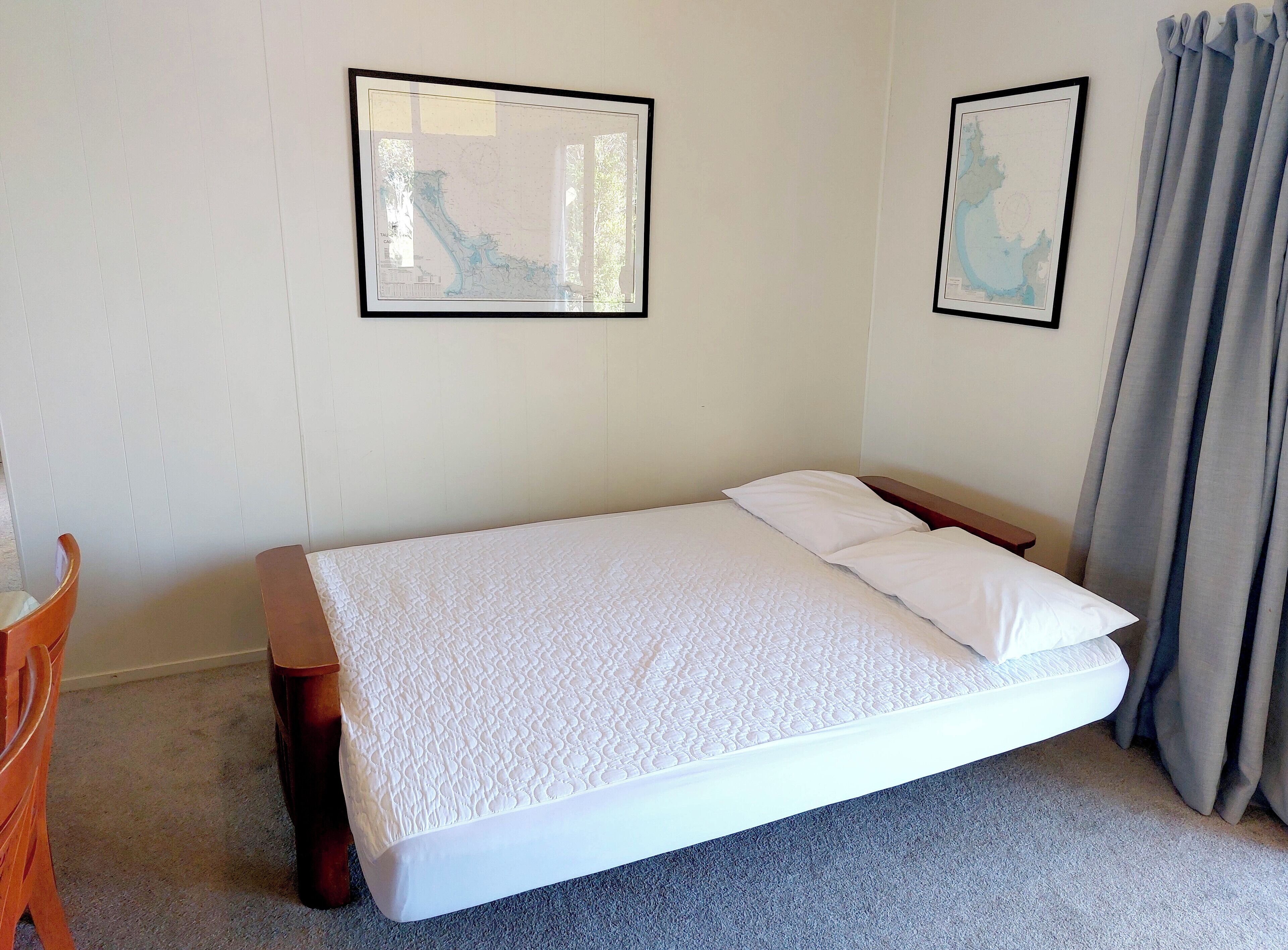 A neatly made bed with a white mattress and pillows, a wooden nightstand, and two framed maps on the wall.