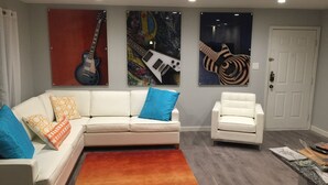 Un salon avec un canapé blanc, un fauteuil blanc et un tapis coloré. Trois guitares sont accrochées au mur.