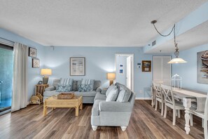 A living room with a sofa, a coffee table, and a dining table with chairs. - Caprice 501 Top Floor Corner|Stunning Beach Views|Walk to everything! (St. Pete Beach)