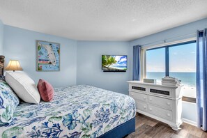 A bedroom with a bed, a dresser, a TV, a map, and a window with a view of the beach.