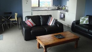 A compact living space with a black leather sofa, a wooden coffee table, and a dining area with a small table and chairs. A kitchen with white cabinets and a stove is visible in the background. - Kākā Point Beach House (Kaka Point)