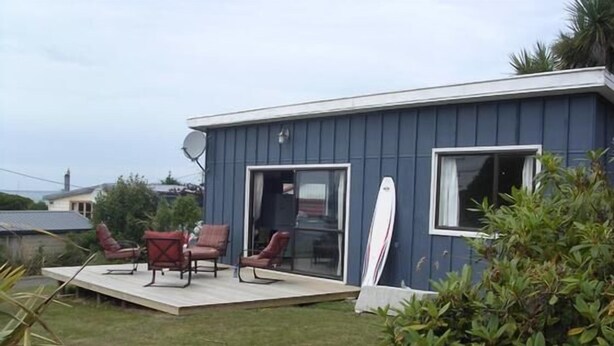 A small house with a patio, a surfboard leaning against the wall, and a satellite dish. - Kākā Point Beach House (Kaka Point)