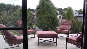 A covered outdoor seating area with four cushioned chairs and a central ottoman, surrounded by greenery. - Kākā Point Beach House (Kaka Point)