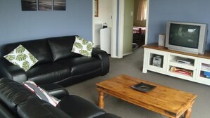 A living room with a black leather sofa, two green leaf-patterned pillows, a wooden coffee table, and a white entertainment unit with a television. - Kākā Point Beach House (Kaka Point)