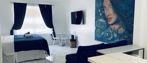A modern hotel room with a bed, a sitting area with a couch and a table, a ceiling fan, a painting on the wall, and a TV mounted on the wall.
