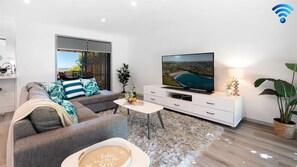 A modern living room with a sofa, a coffee table, a flat-screen TV, and a view of the sea. - Heights Hideaway – breathtaking coastal and rural views (Kiama Heights)
