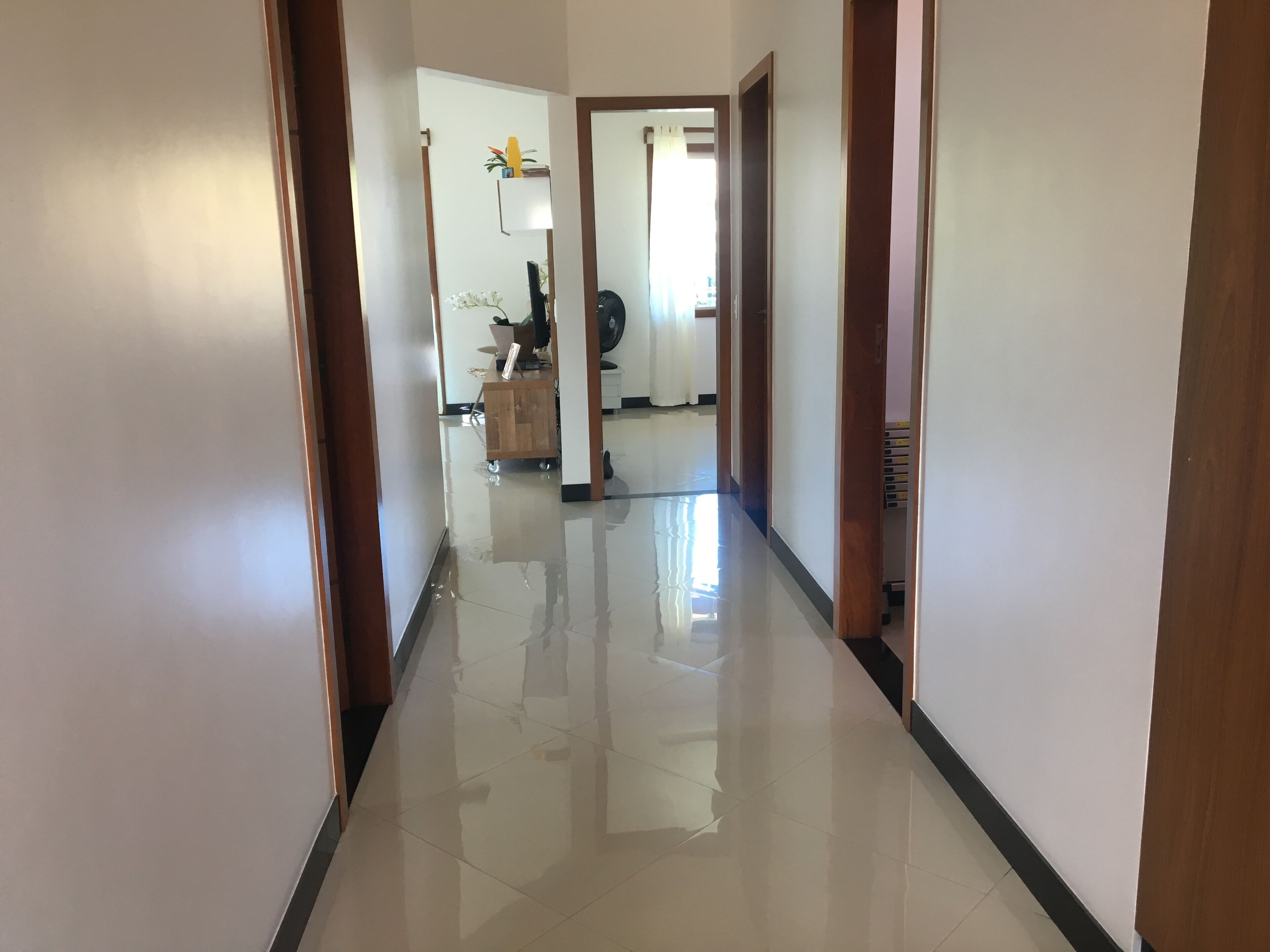 A hallway with a shiny tiled floor, wooden doors, and a small wooden cabinet with a fan and decorative items.