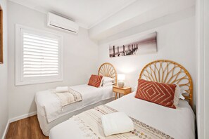 Habitación de hotel con dos camas, una ventana con persianas, un equipo de aire acondicionado, una lámpara de noche y un cuadro colgado en la pared.