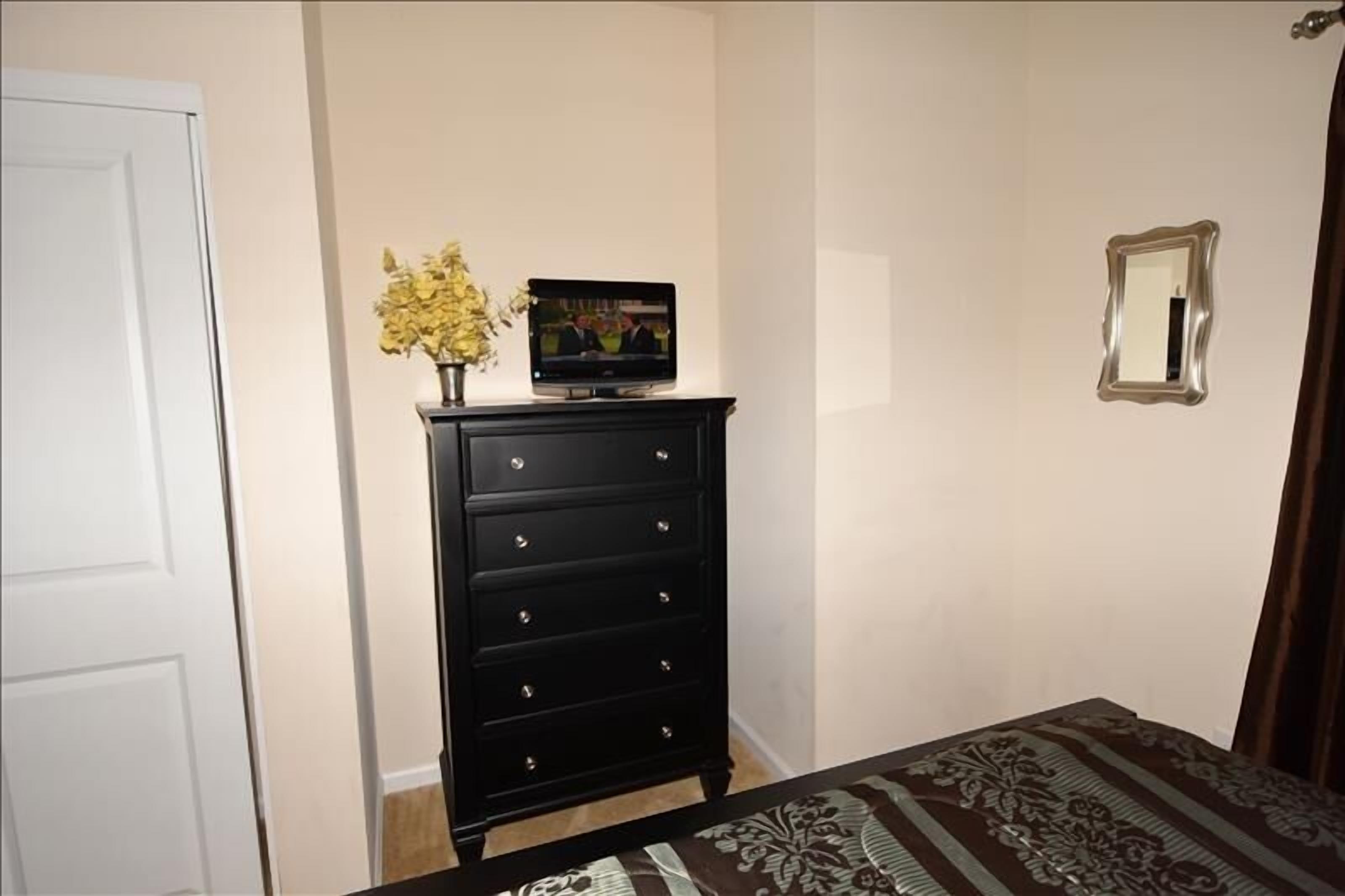 Une chambre à coucher avec un lit, une commode, un téléviseur posé sur la commode, un miroir au mur et une porte.