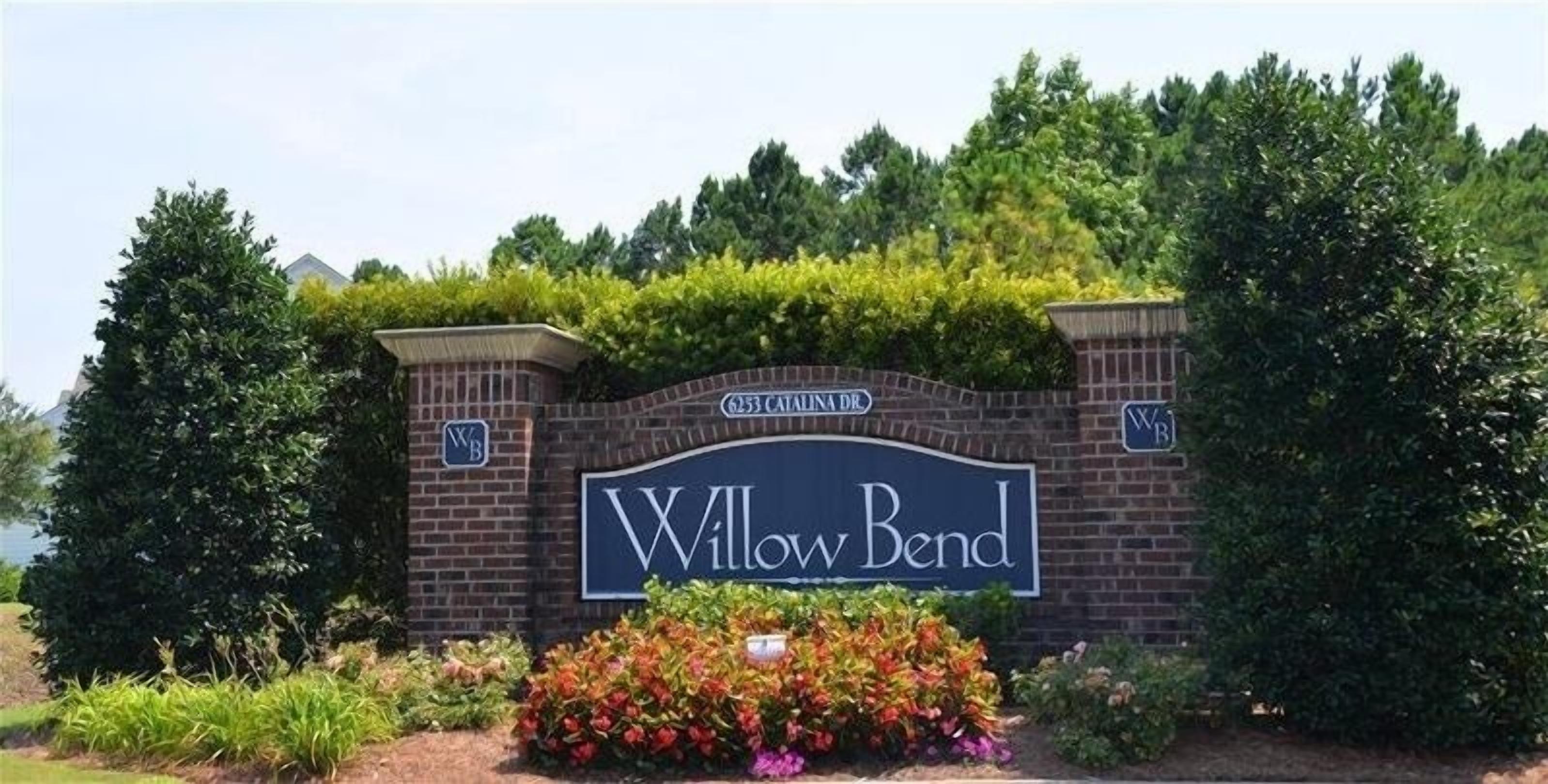 A sign for Willow Bend with a brick wall and greenery.