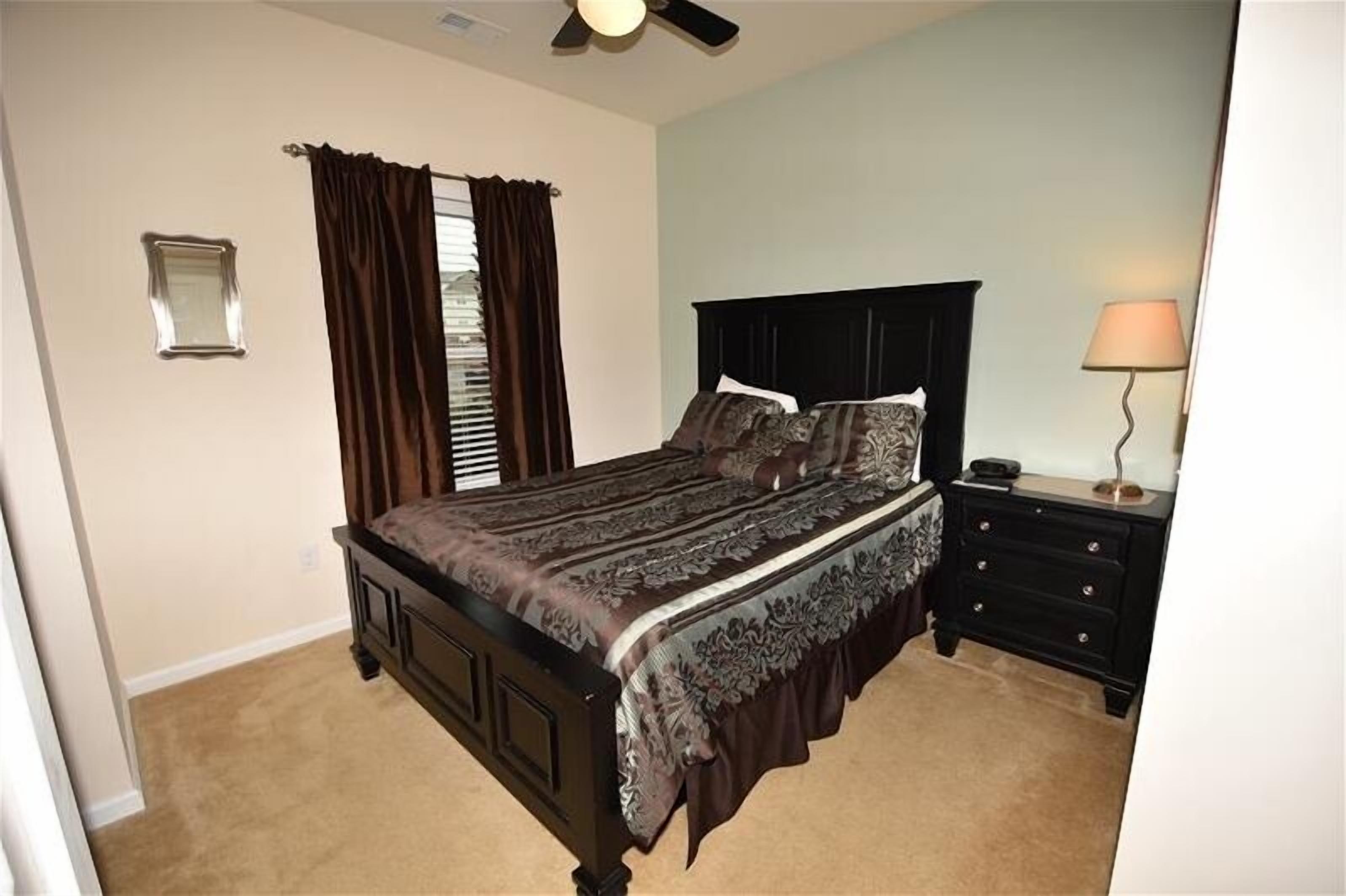 A bedroom with a bed, a nightstand, a lamp, a ceiling fan, and a window with curtains.