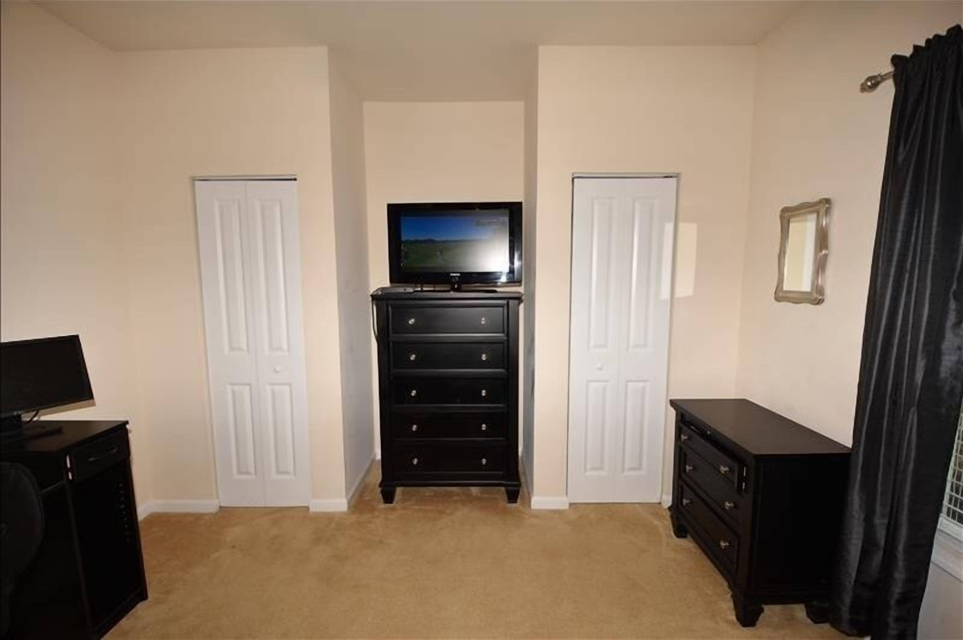 A room with beige carpet, white doors, a black dresser, a television on a stand, and a framed picture on the wall.