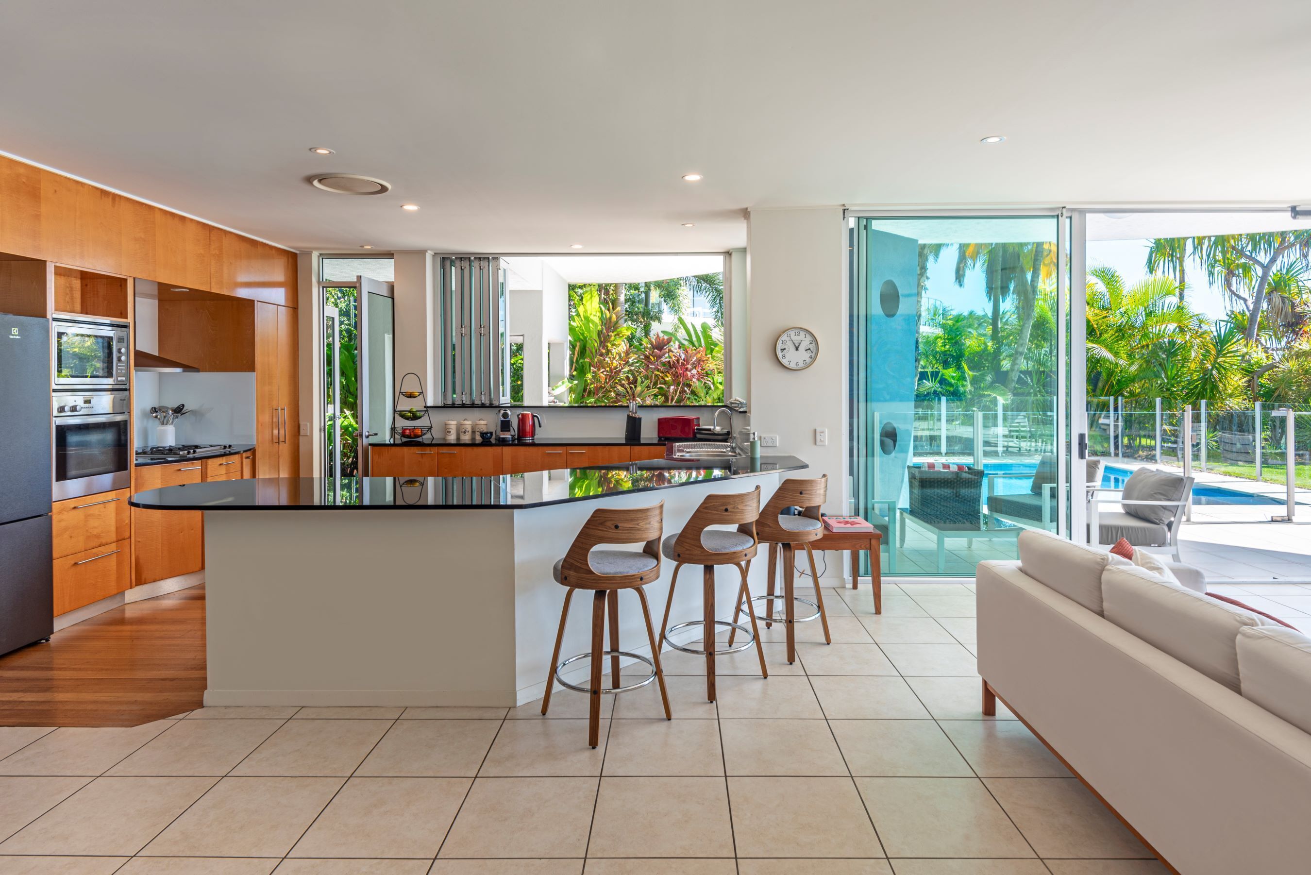 A modern kitchen with a central island, bar stools, and a view of a pool and greenery.