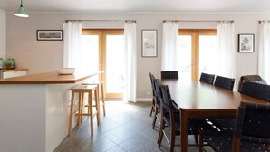 A dining area with a wooden table and chairs, a kitchen counter with a bowl, and a hanging light fixture. - Solomon Street Cottage (Fremantle)