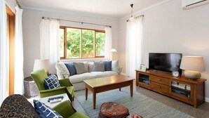 A living room with a sofa, green armchair, wooden coffee table, TV, and a side table with a lamp. - Solomon Street Cottage (Fremantle)