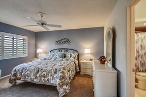 A bedroom with a bed, two bedside tables with lamps, a ceiling fan, a large mirror, and a window with blinds.