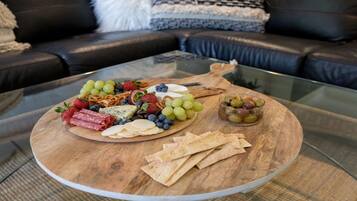 Une planche en bois, sur laquelle sont disposés divers en-cas et des fruits, est posée sur une table en verre dans un salon.