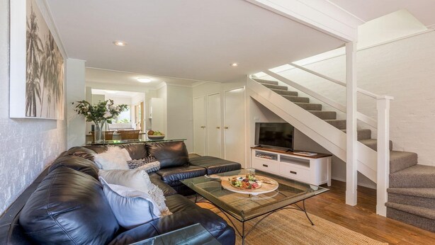 Un salon moderne avec un canapé en cuir, une table basse en verre et un téléviseur à écran plat posé sur un meuble de télévision blanc. Un escalier au revêtement en bois mène à l’étage supérieur.
