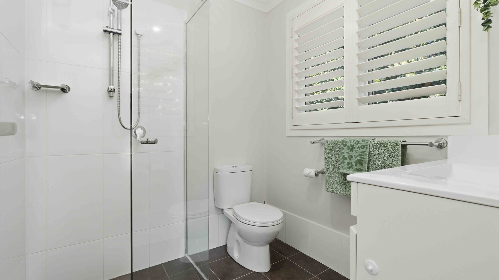 A bathroom with a glass shower enclosure, a white toilet, a window with blinds, and a white sink.