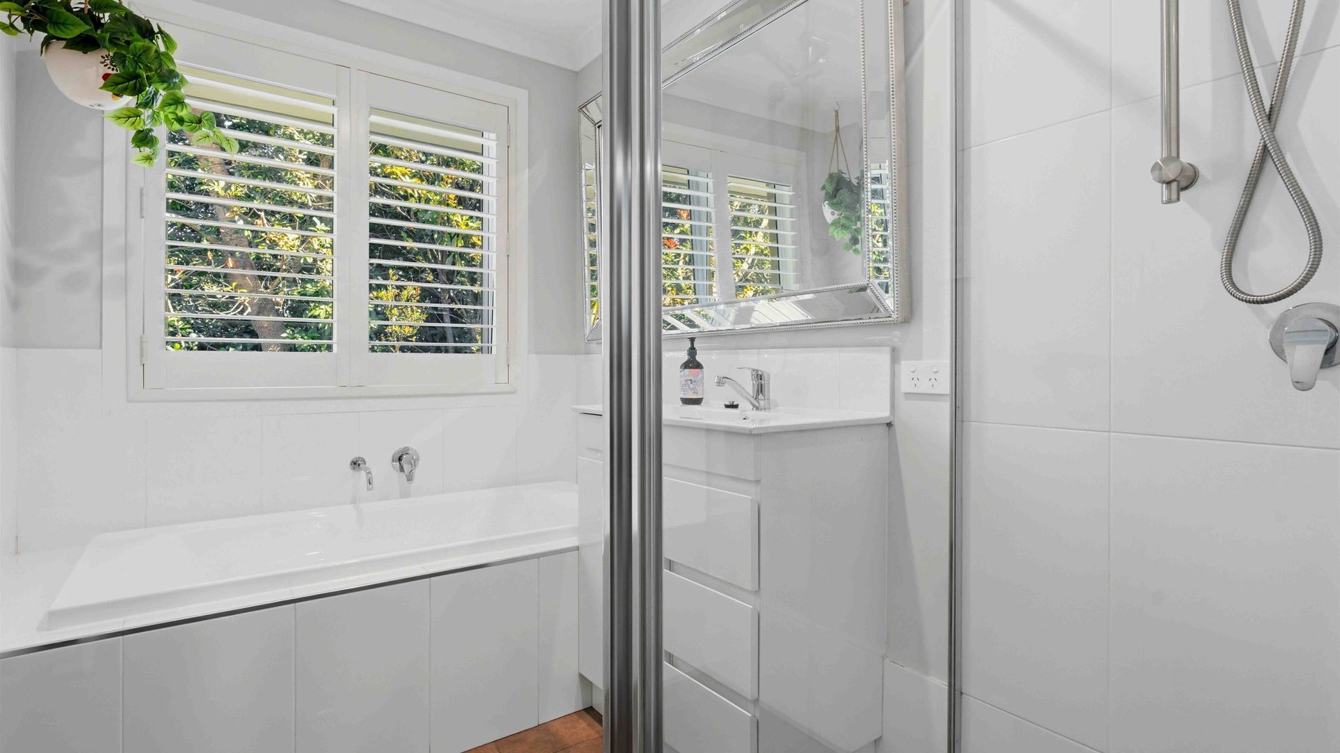 A bathroom with a glass shower enclosure, a white sink, and a window with blinds.