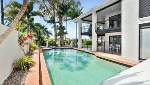 Un área de piscina moderna con una piscina cristalina, rodeada de palmeras y un edificio de dos pisos con balcones.