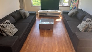 Ein Wohnzimmer mit zwei dunklen Sofas, einem Glastisch und einem Flachbildfernseher auf einem Standfuß.