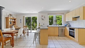 A modern kitchen with a central island, a dining area with a table and chairs, and a view of greenery outside. - Husky House by Experience Jervis Bay (Huskisson)