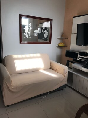 A living room with a beige sofa, a framed artwork, a television, and a small shelf with decorative items.