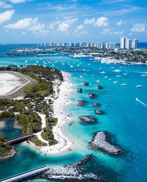Une vue aérienne d’une zone côtière avec une plage, des bateaux et, au loin, le profil urbain d’une ville.