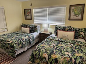 Une chambre à coucher avec deux lits, chacun agrémenté de draps à motifs floraux, une table de chevet, une lampe et une fenêtre avec des stores.