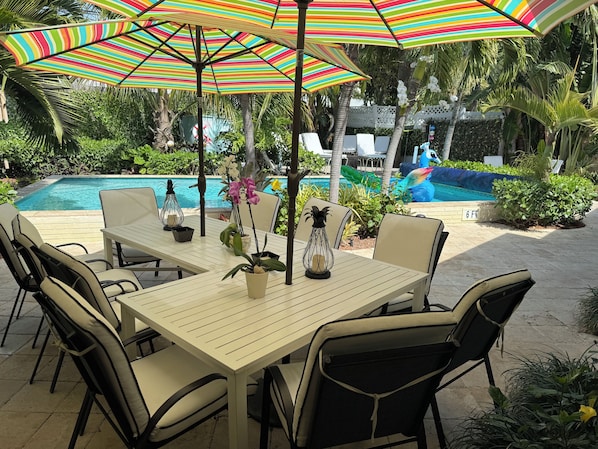 Un espace aménagé au bord de la piscine, avec une table et des chaises, un parasol rayé aux couleurs vives, et une piscine en arrière-plan.