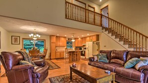 A spacious living room with leather sofas, a wooden coffee table, and a large rug. A staircase leads to an upper level, and a kitchen is visible in the background. - Pocono Mountain Home w/ 3 Community Lakes! (Gouldsboro)
