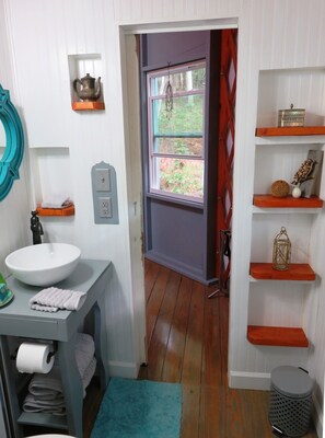 A bathroom with a white sink, a mirror, a towel, a shelf with decorative items, and a window with a view of greenery. - Unique and Charming BlueBird Yurt at Feathered Fox Farm (Rutherfordton)