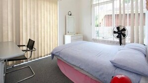 A bedroom with a bed, a desk, a chair, a dresser, and a window with blinds.