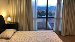 Une chambre d’hôtel avec un grand lit, une table de chevet avec une lampe et une vue sur le paysage urbain, visible à travers une fenêtre avec des rideaux.