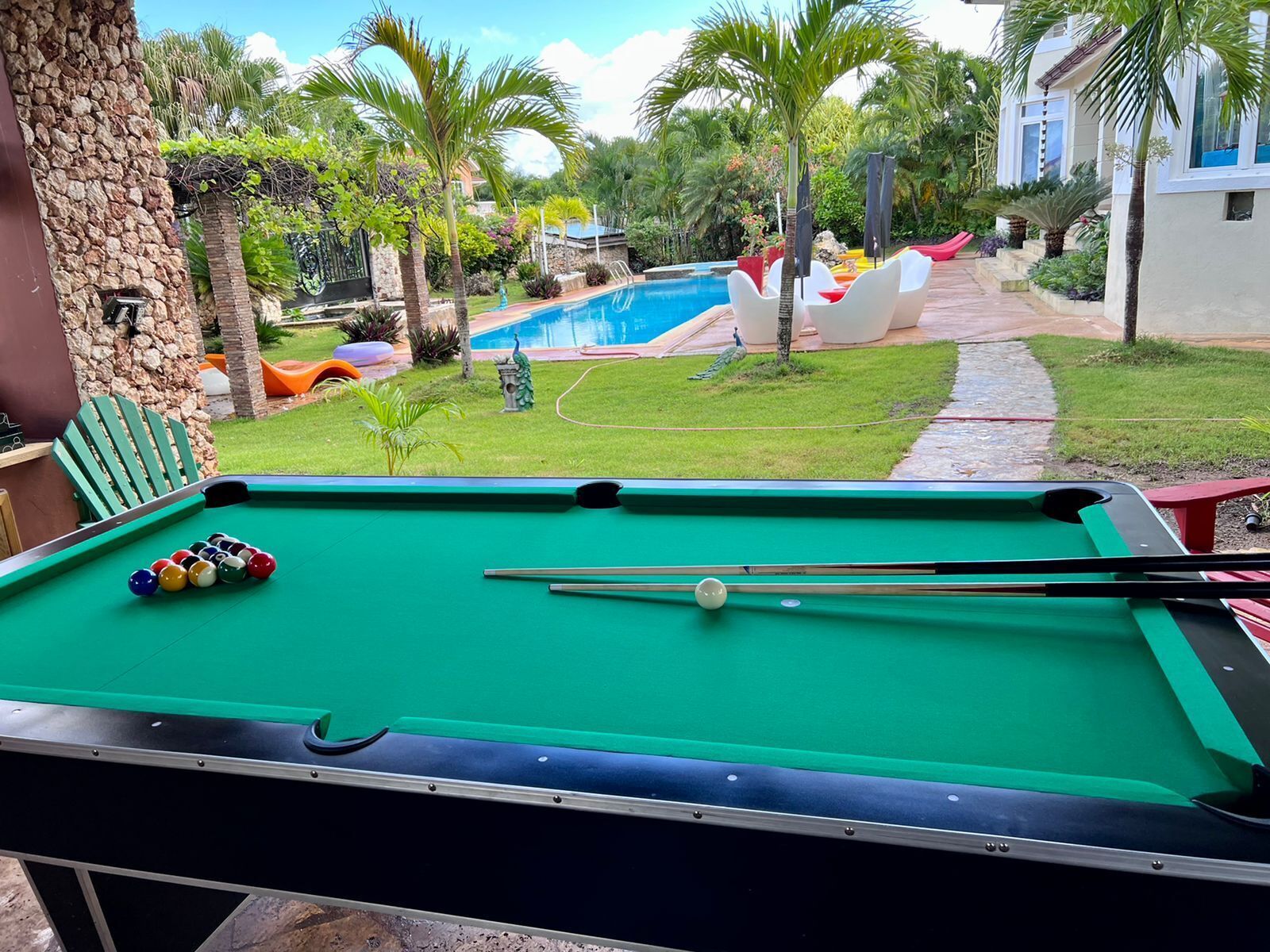 Ein Billardtisch mit Kugeln und Spielstöcken, eine Steinwand und Blick auf einen Swimmingpool und Grünflächen.