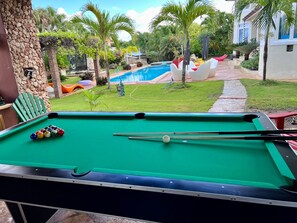 A pool table with balls and cues, a stone wall, and a view of a swimming pool and greenery. - Tropical Villa Ideal for Family Gatherings (Sosua)