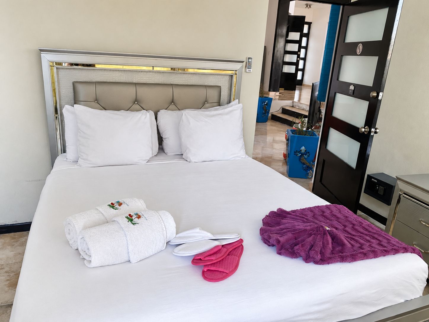 Una cama bien hecha con sábanas blancas, una colcha morada y toallas enrolladas.