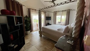Une chambre spacieuse avec un grand lit, un ventilateur de plafond, une télévision et un balcon agrémenté de rideaux.