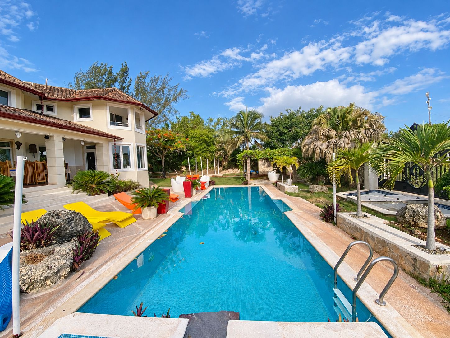 A swimming pool with a stone deck, surrounded by tropical plants and a two-story building with a tiled roof.
