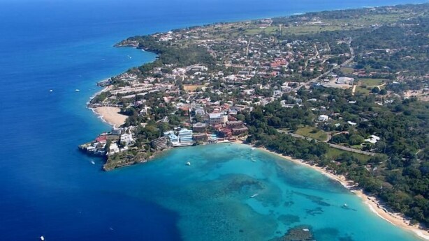 Vue aérienne d’une ville côtière avec une plage, des eaux cristallines et des bâtiments.