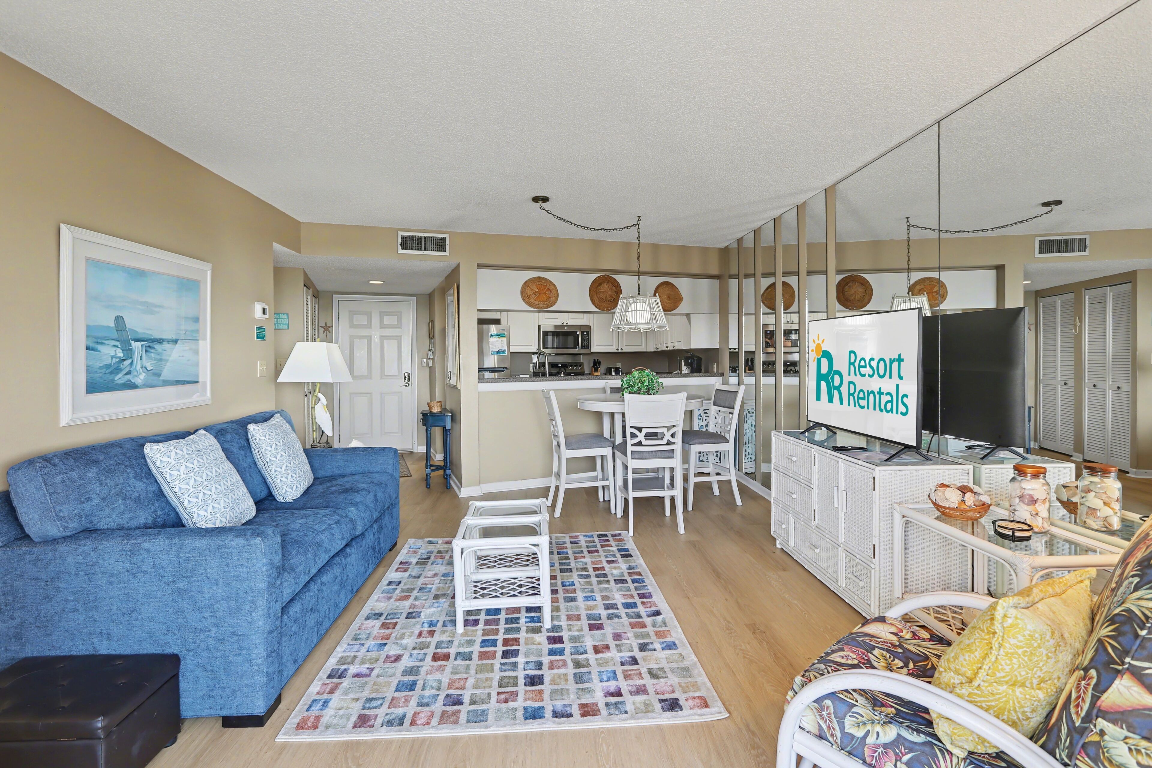 A modern living room with a blue sofa, a patterned rug, and a dining area with white furniture. A sign for 'Resort Rentals' is visible.